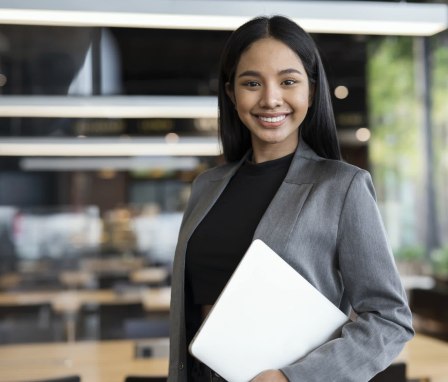 Smart People with Technologies to Boost Business Success. Portrait of Confident Young Female Entrepreneur standing while holding a laptop in a modern business office.