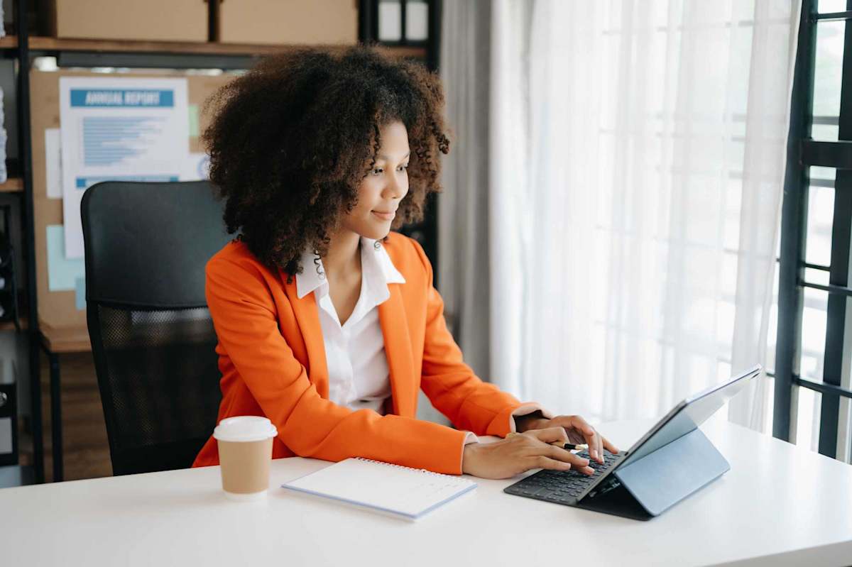 Young African woman typing on tablet and laptop while sitting at the working wooden table modern office