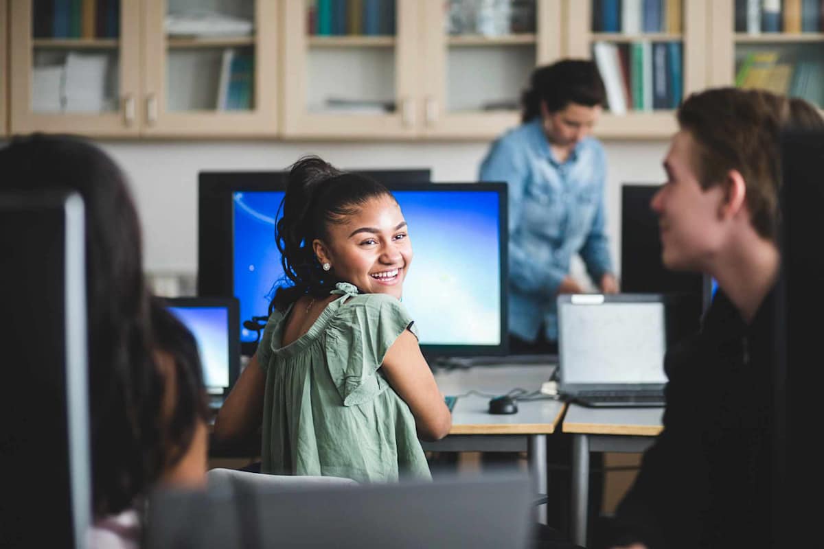 Smiling female high school student looking at friend sitting in computer lab