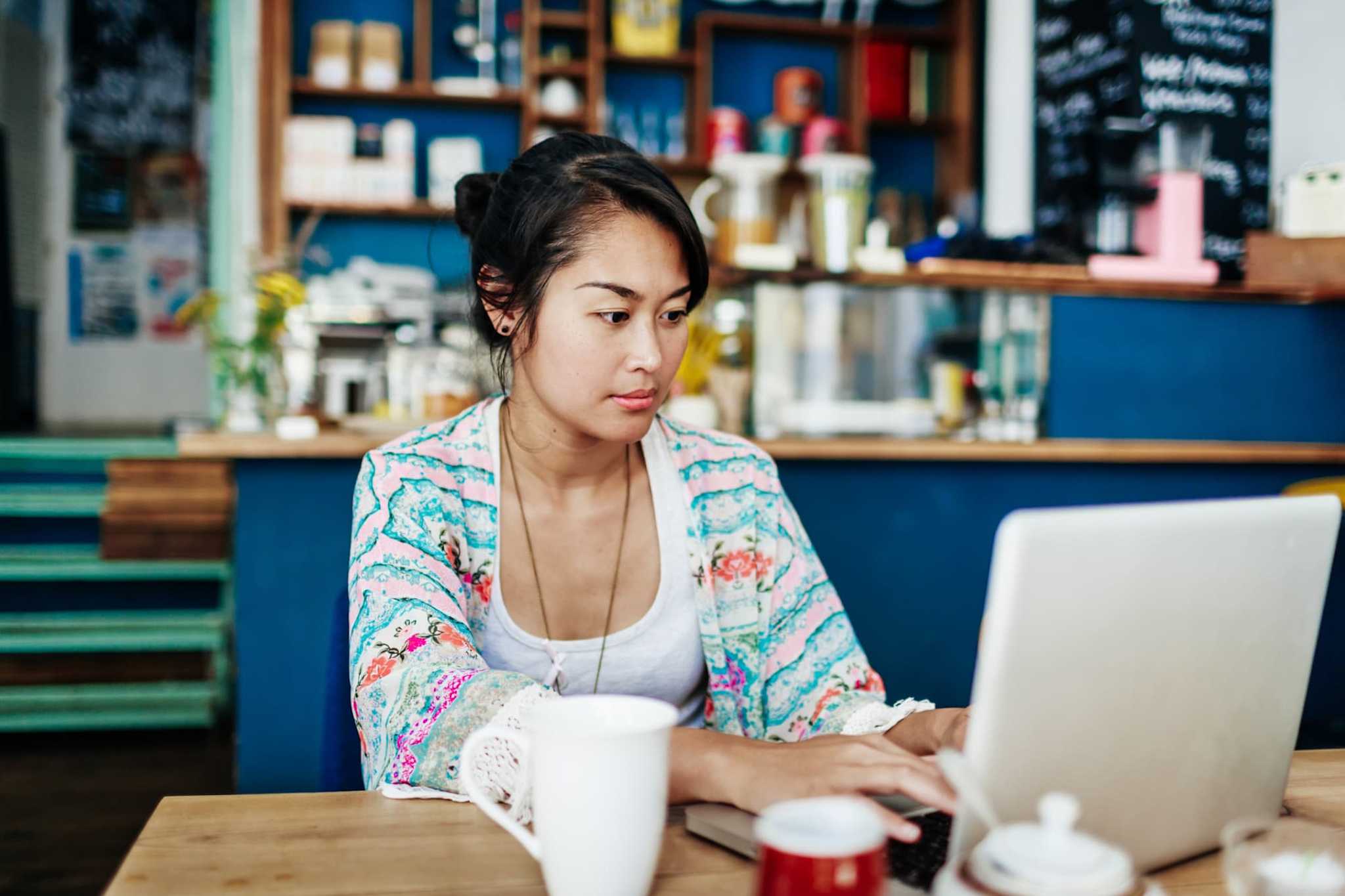 A female Asian American coding bootcamp student sitting at a table in a cafe. She is working on an assignment for class on her laptop computer.