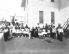 Black and white photo of an old building with a group of Black children and their teacher standing outside of it.