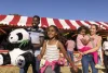 A family enjoying a day out at the fair. They are walking through the fairground away from a fairground stall while smiling. The father is carrying a plush toy panda that they have won at the fair and the mother is carrying the youngest daughter.