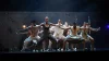 A group of dancers holding wide open arms and leg poses in a black box theater setting