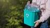 A person carries a recycling container full of empty, rinsed, plastic bottles with no lids.