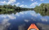 The front of an orange kayak looking out over the river at Camp Bayou with a bright blue sky filled with clouds and green trees all along the river banks