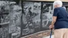 An older woman with a cane stands in front a memorial wall that shows pictures from war scenes