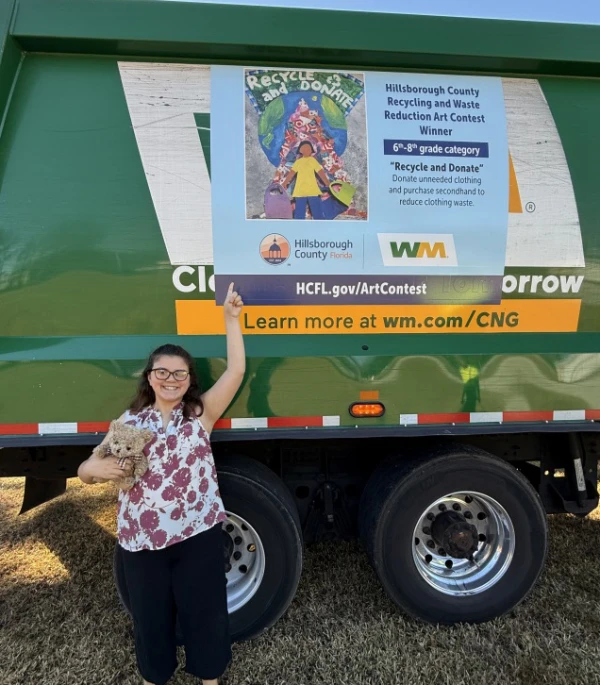 A girl holding a teddy bear in front of green recycling truck points to her winning artwork on the side: "Recycle and Donate" - Donate unneeded clothing and purchase second hand to reduce clothing waste.