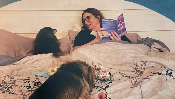 Lady in her bed with her two dogs holding a book and looking at one of the dogs