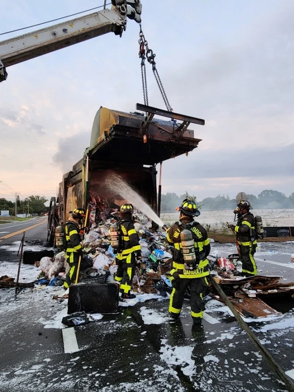 Firefighters in full gear spraying water into the back of a garbage truck that has an active fire going on.