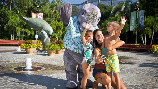 The Manny-T mascot in a Hawaiian shirt covered in manatees poses with a mom and her two young kids in front of a manatee sculpture