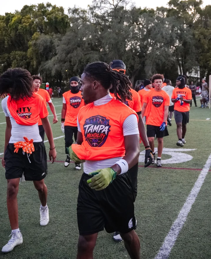 A group of young men in bright orange Tampa Mentality shirts are on a football field