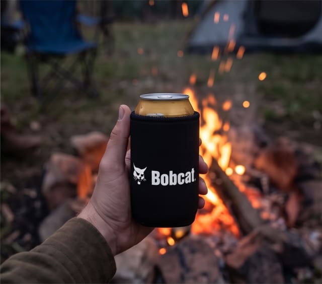 Hand holding a drink in a Bobcat stubby holder in front of a campfire