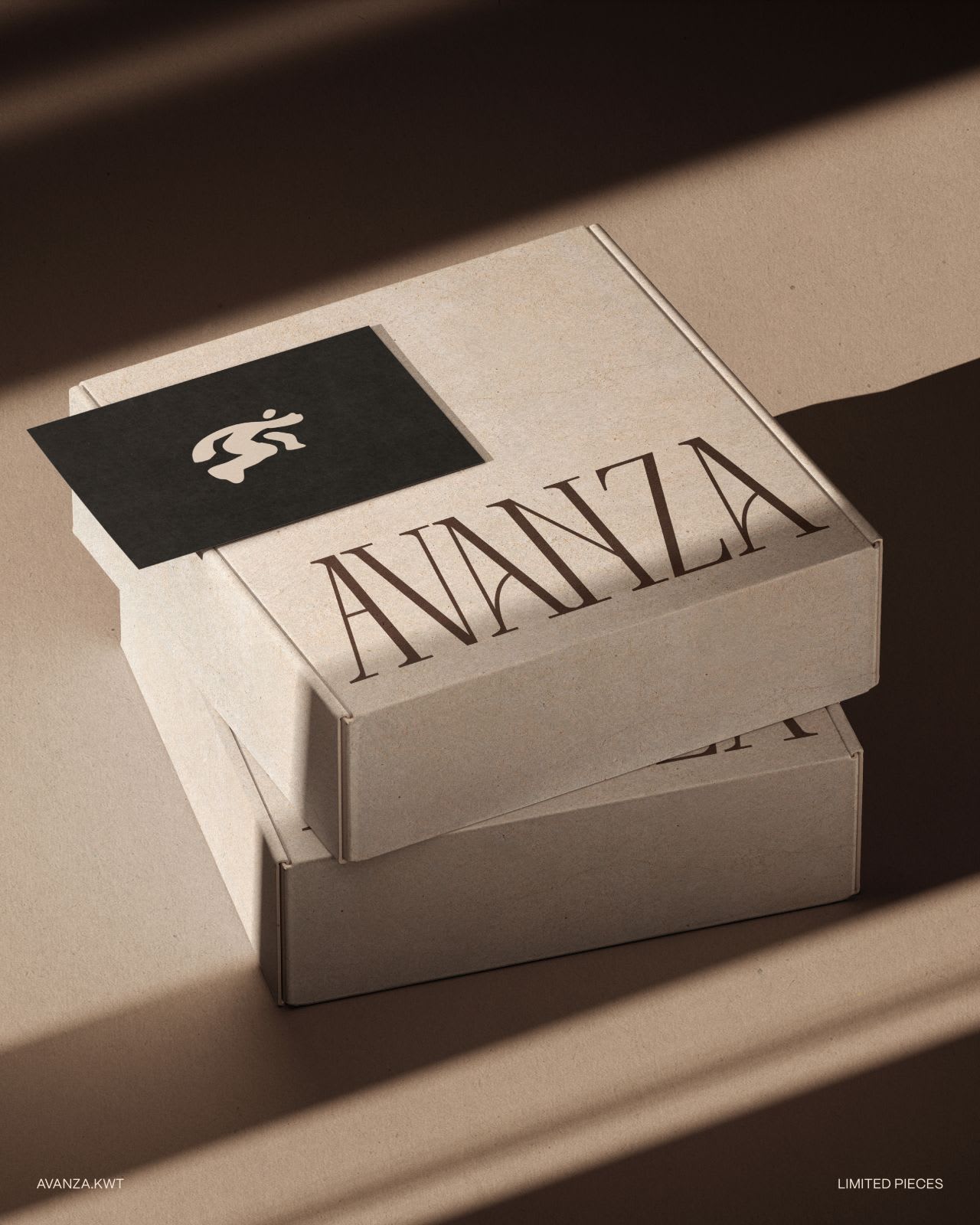 Two stacked kraft mailer boxes printed with the word AVANZA in a high-contrast serif, a charcoal business card resting on the lid, hard diagonal window light across a warm beige surface.