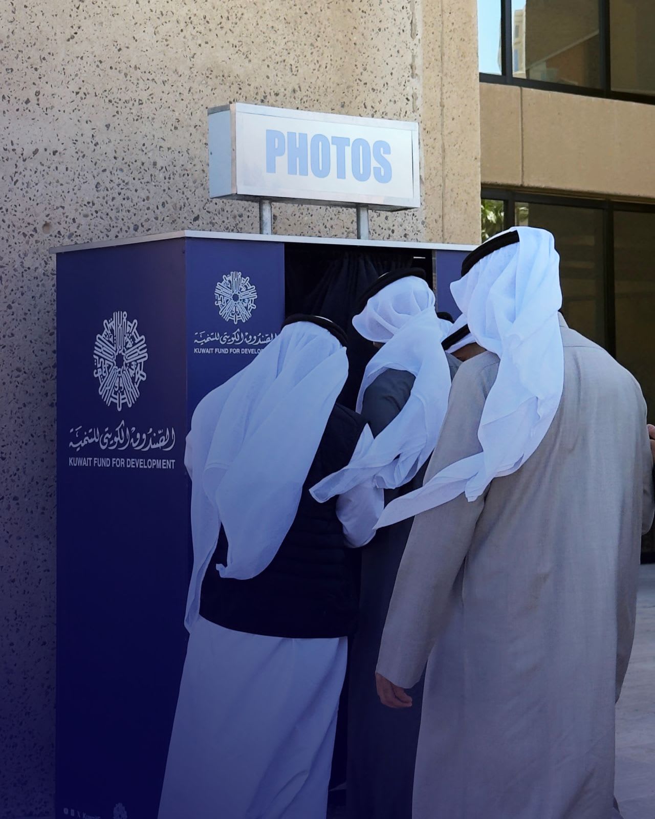 Three people in white Kuwaiti dishdasha and ghutra gathered around a purple branded photo booth marked PHOTOS, outdoors beside a stone building.
