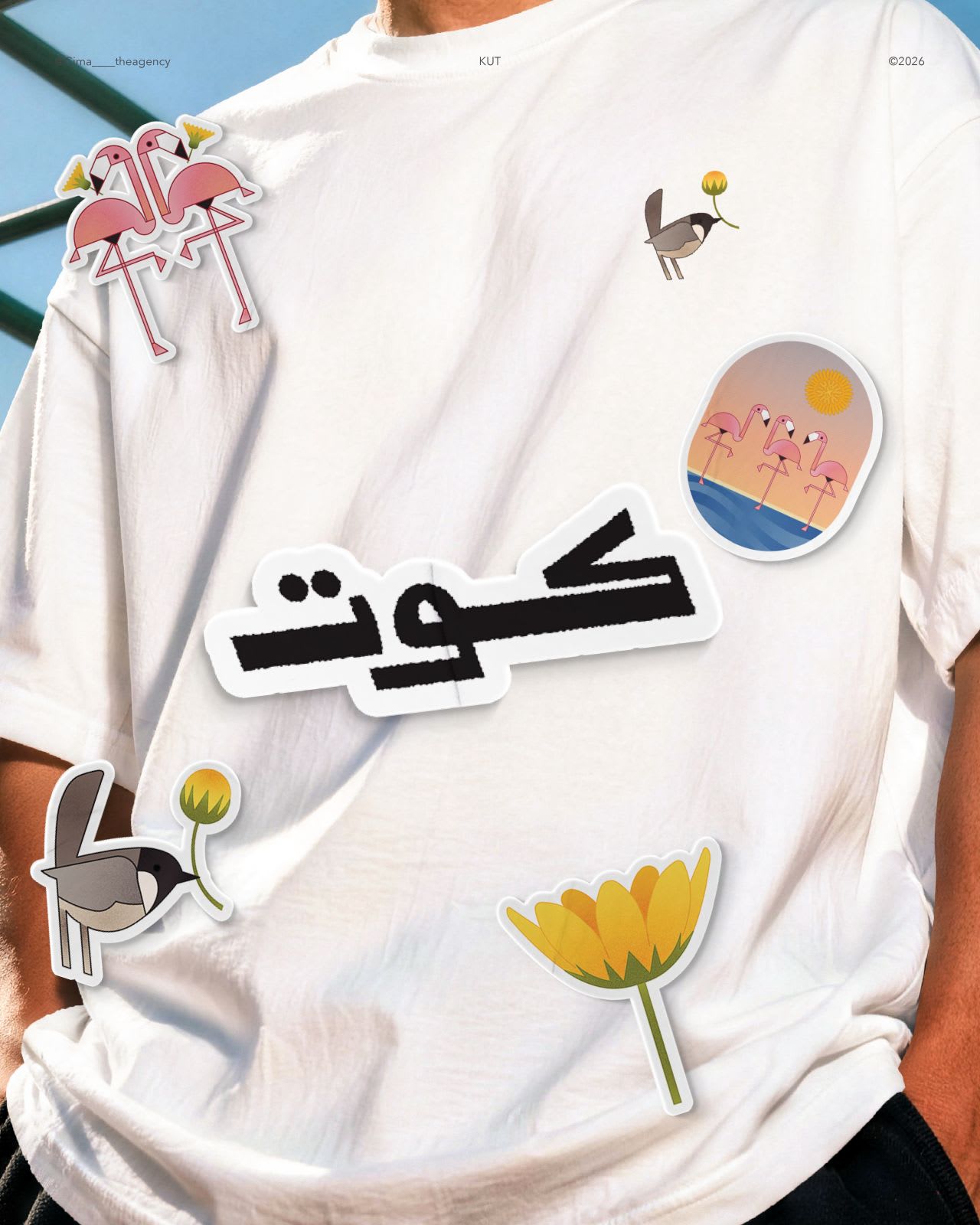 A person wearing an oversized white T-shirt against a blue sky, decorated with illustrated stickers of pink flamingos, a small bird holding a yellow flower, and the word Kuwait in Arabic script.