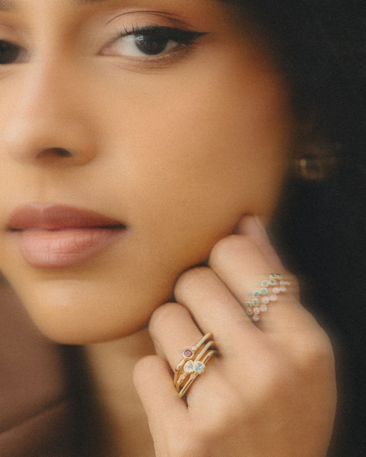 A close-up portrait, shallow focus, of a woman resting her chin on her hand and looking off camera, wearing several slim gold rings set with pale and green stones.