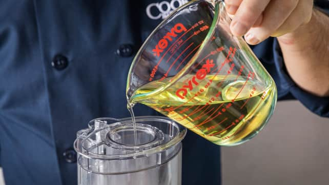 A hand drizzling a measuring cup of oil into the top of a food processor.