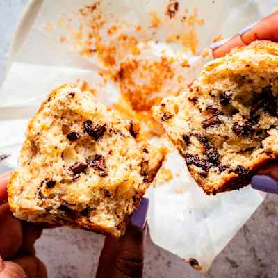 Two hands with lavender-painted fingernails hold a split Browned Butter Chocolate Chunk Muffin
