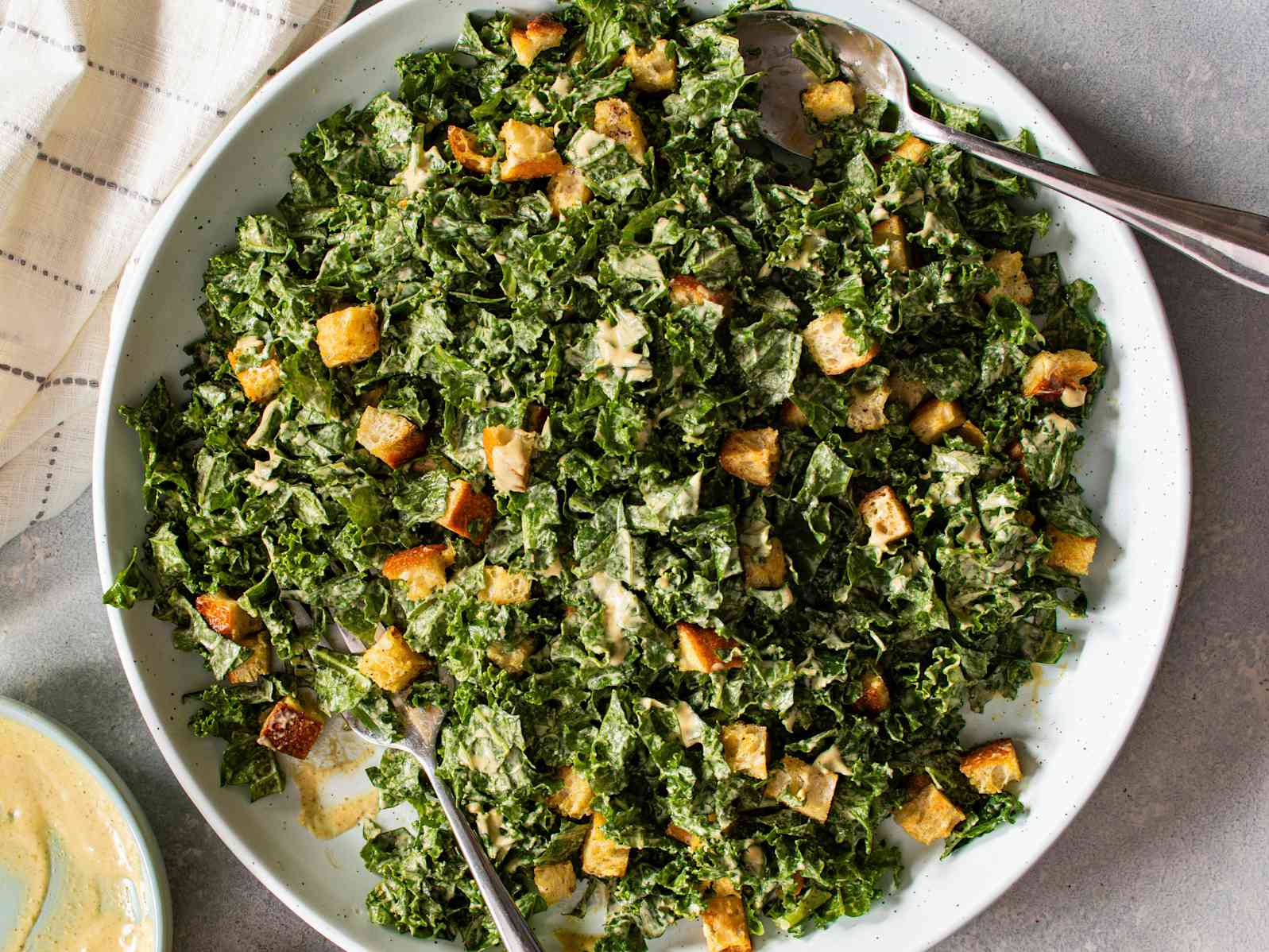Kale Caesar Salad in a large white bowl with two spoons for serving, on a gray table with a napkin and a dish of extra dressing seen in the bottom left corner