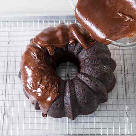 Chocolate glaze being poured over the chocolate Bundt pan on a wire rack set inside a rimmed baking sheet.