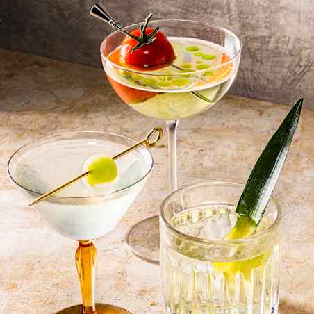 A coupe glass with a clear drink and a tomato garnish, a martini glass with a pickled-onion garnish, and an old-fashioned glass with an aloe-leaf garnish, all sitting on a brown surface.