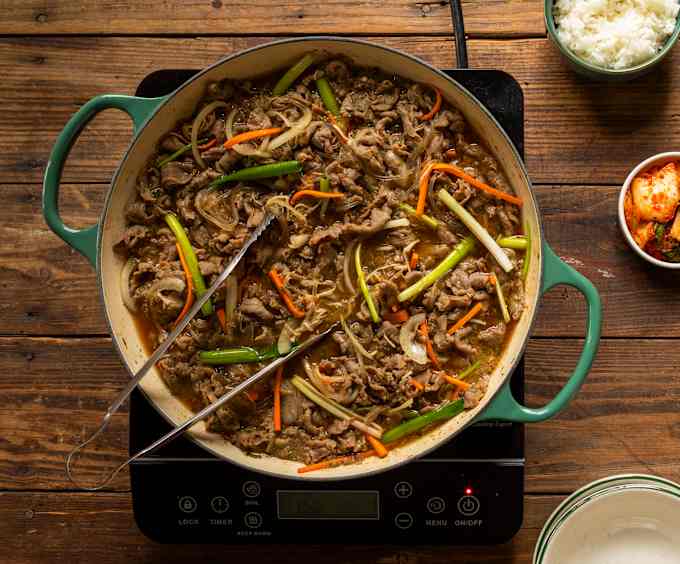 A green braiser sits on top of an induction cooktop. It contains thinly sliced meat, green onions, and carrots. 