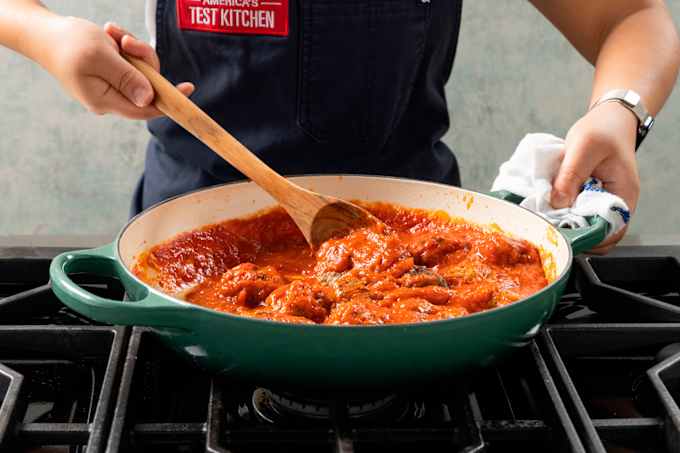 A green braiser full of meatballs and marinara sits on top of a range. One hand reaches in with a wooden spoon to stir the sauce.