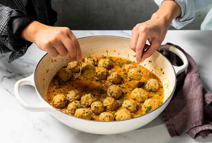 Hands reach in with toothpicks to spear Spanish-style meatballs served in a white braiser.