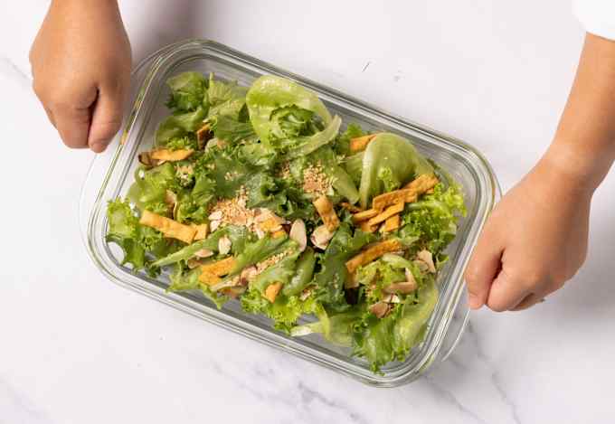 A person grasps the wide rims on the ends of a glass container of green salad.
