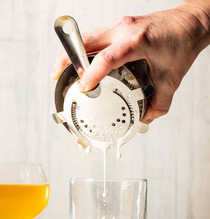 A hand uses a Hawthorne strainer with a metal cocktail shaker to pour out a drink.