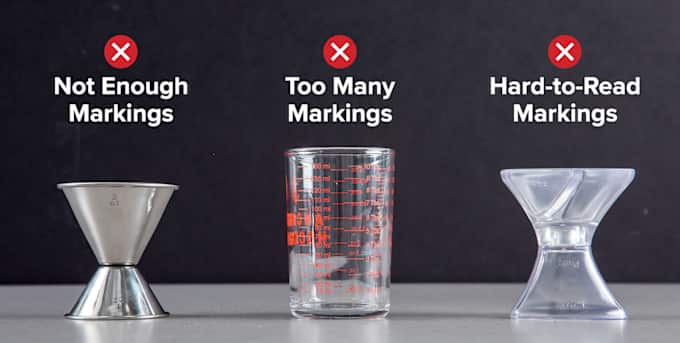 Three jiggers sit on a grey surface. The one on the left is a metal jigger and has just one etched measurement marking. The one in the middle is glass and has many red measurement markings. And the one on the right is made of translucent plastic with hard-to-read etchings.
