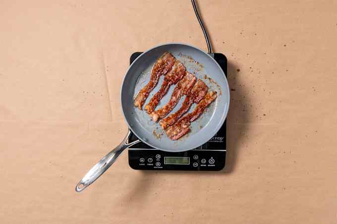 Five strips of cooked bacon sit inside a skillet on a portable induction cooktop. The cooktop is on top of brown kraft paper with visible grease splatter. 