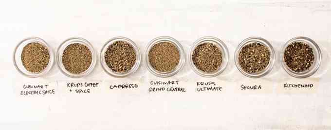 On a white background, glass bowls of ground peppercorn made using 7 different grinders (labeled) line up to show the different consistencies of each grinder. 