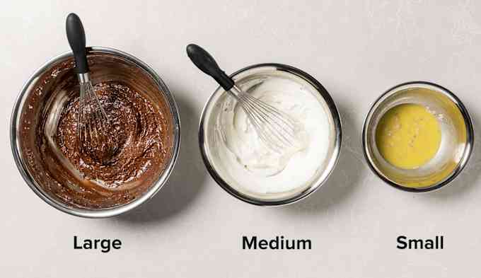 A large stainless steel bowl with brownie batter, a medium stainless steel bowl with whipped cream, and a small stainless steel bowl with vinaigrette.