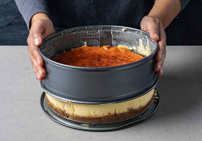The loosened collar of a springform pan is lifted off of a cheesecake.