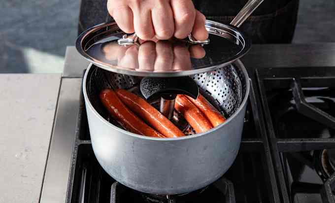 a person lifts the lid of a saucepan, exposing the four hotdogs in a collapsible steamer basket that’s nestled inside the pot.