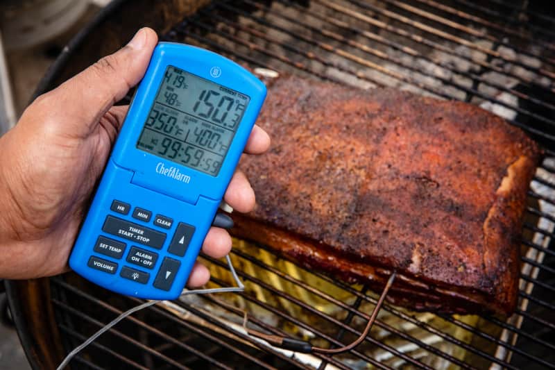 Smoked pork slab on a charcoal grill with a thermometer in it reading 150 degrees.