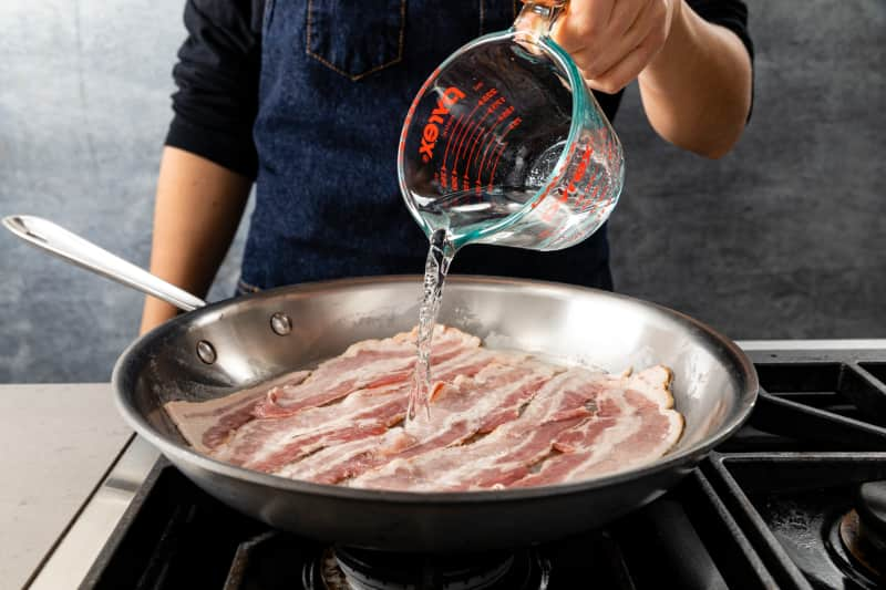 pouring water from liquid measuring cup into skillet full of bacon