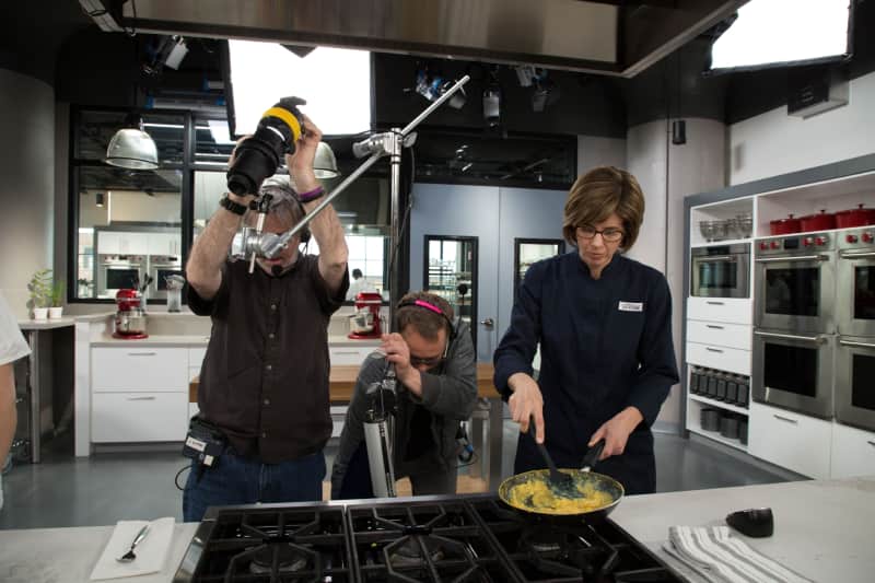 Onscreen test cook and Cook's Illustrated deputy editor Becky Hays continues scrambling eggs for a Creamy French-Style Scrambled Eggs recipe segment while two members of the film crew set up for the perfect shot.