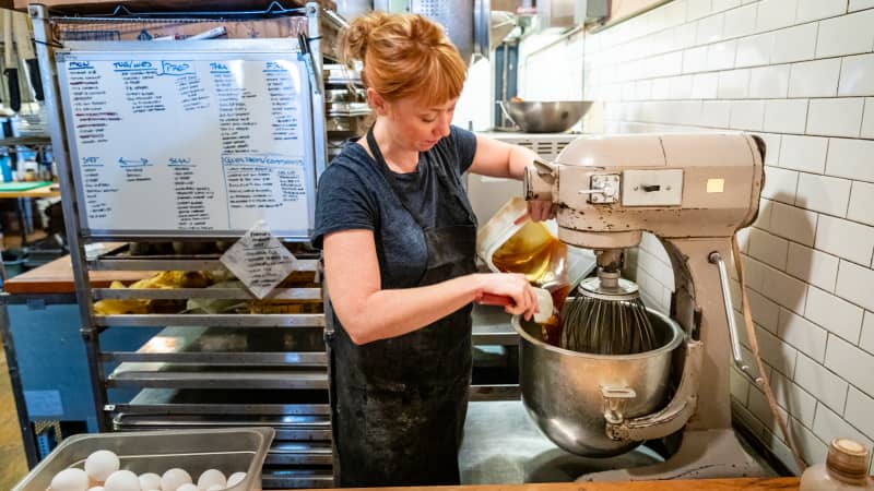 Owner Kate McMillen pours honey from a Cambro into the bowl of a stand mixer