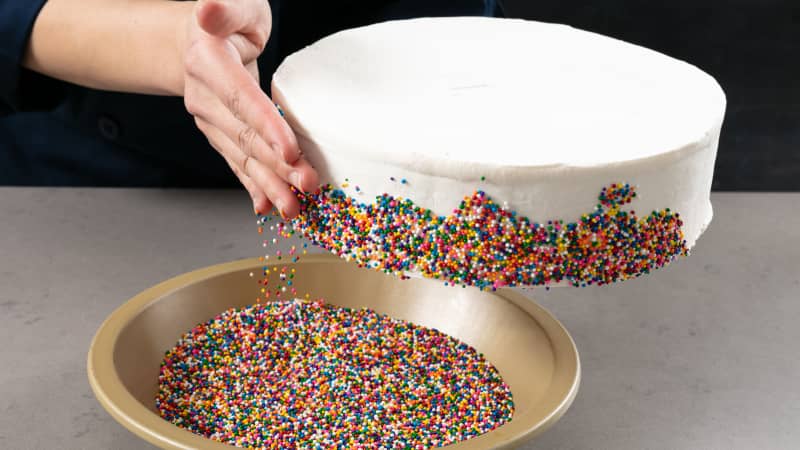 Rainbow pearl sprinkles being pressed onto the side of the ice cream cake.