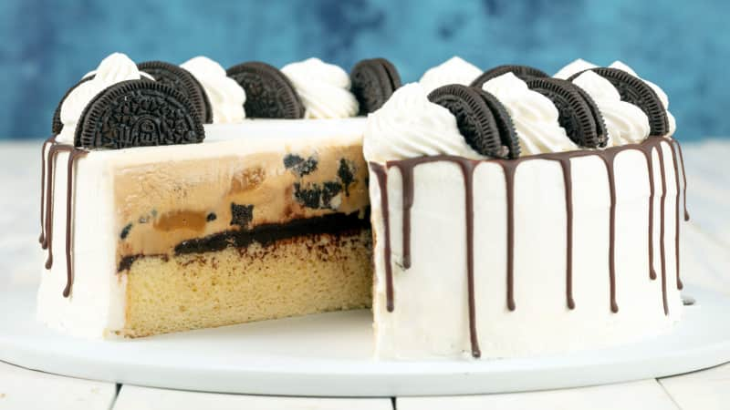 An oreo-decorated ice cream cake with a slice cut out of it showing the fudgy center with an ice cream layer and a cake layer.