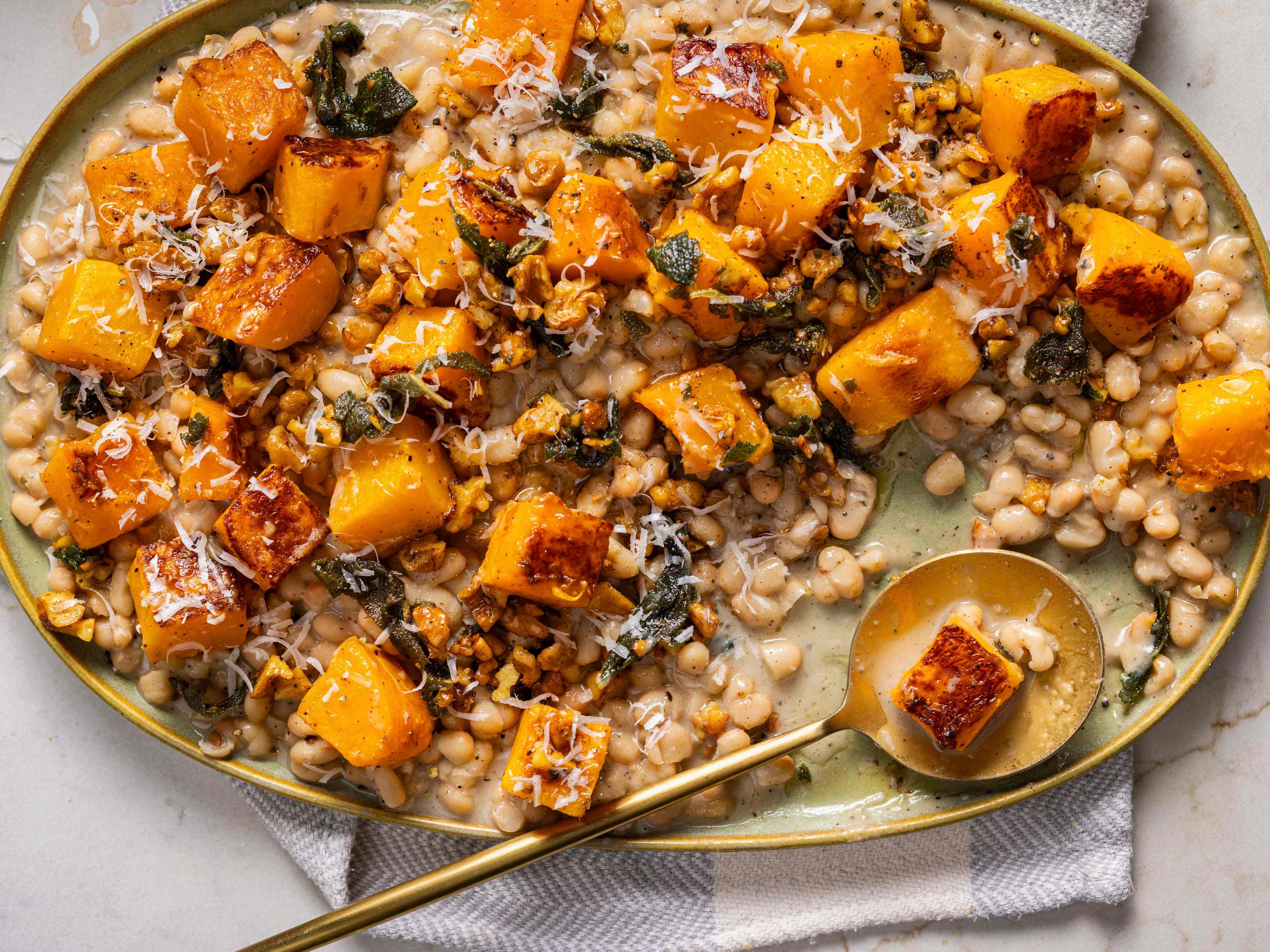 Cacio e Pepe Beans with Squash, Sage, and Walnuts on an oval-shaped serving dish with a round golden spoon