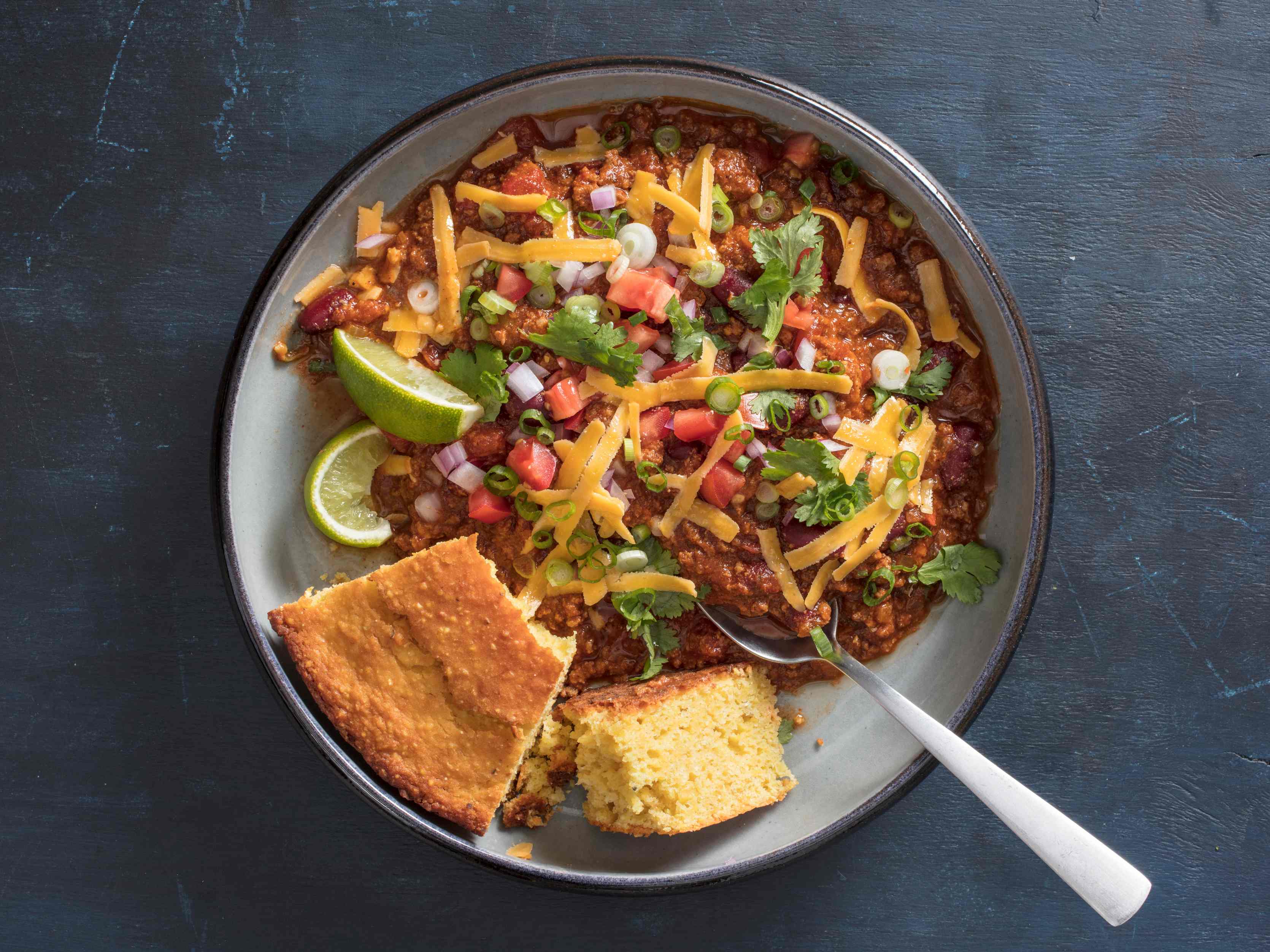 Slow-Cooker Classic Beef Chili garnished with lime wedges, grated yellow cheddar cheese, cilantro, fresh tomato, and scallions serve in a bowl with a wedge of cornbread and a spoon