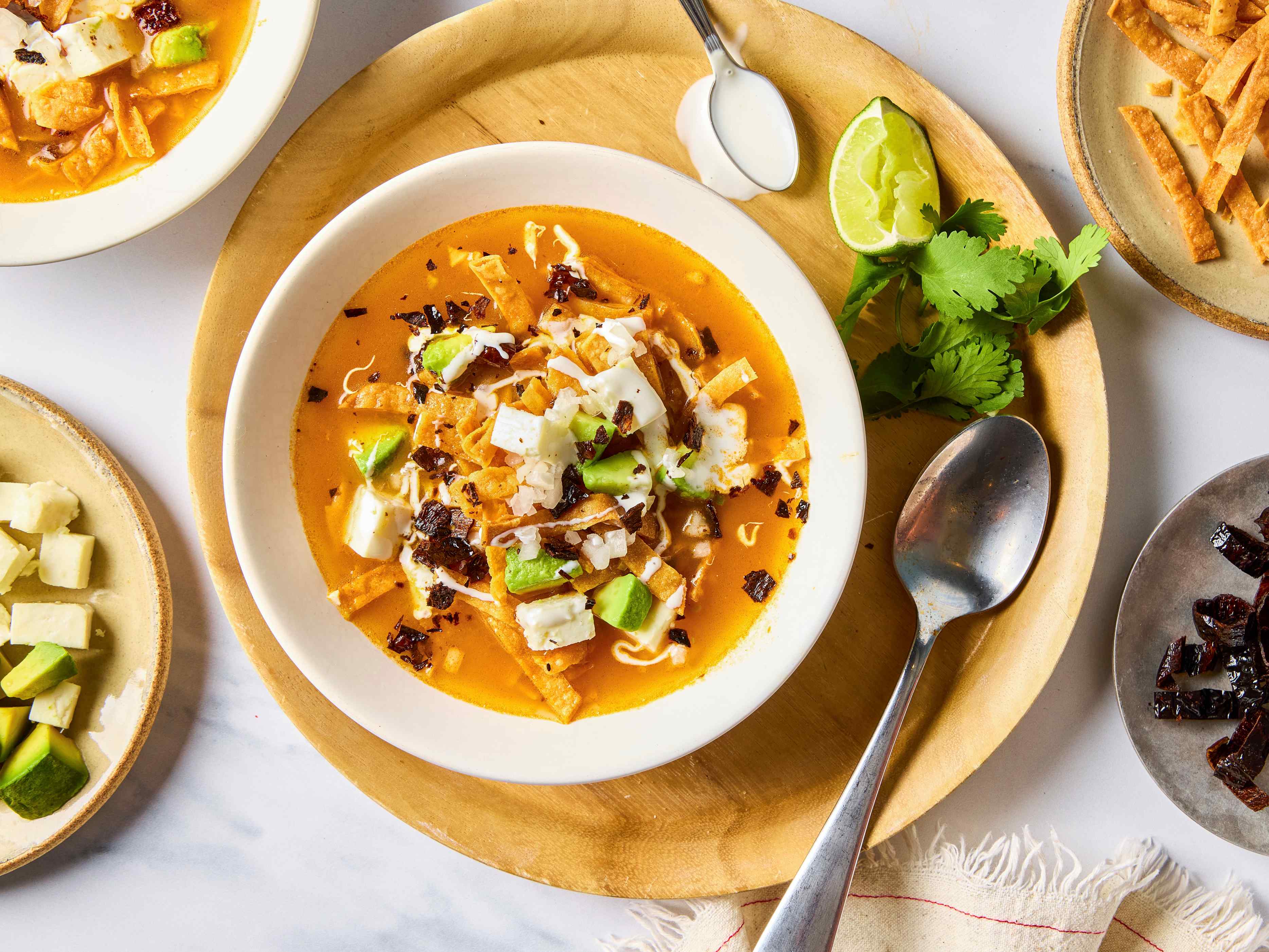A bowl of sopa de tortilla on a round wooden tray with a spoon, surrounded by dishes of garnishes