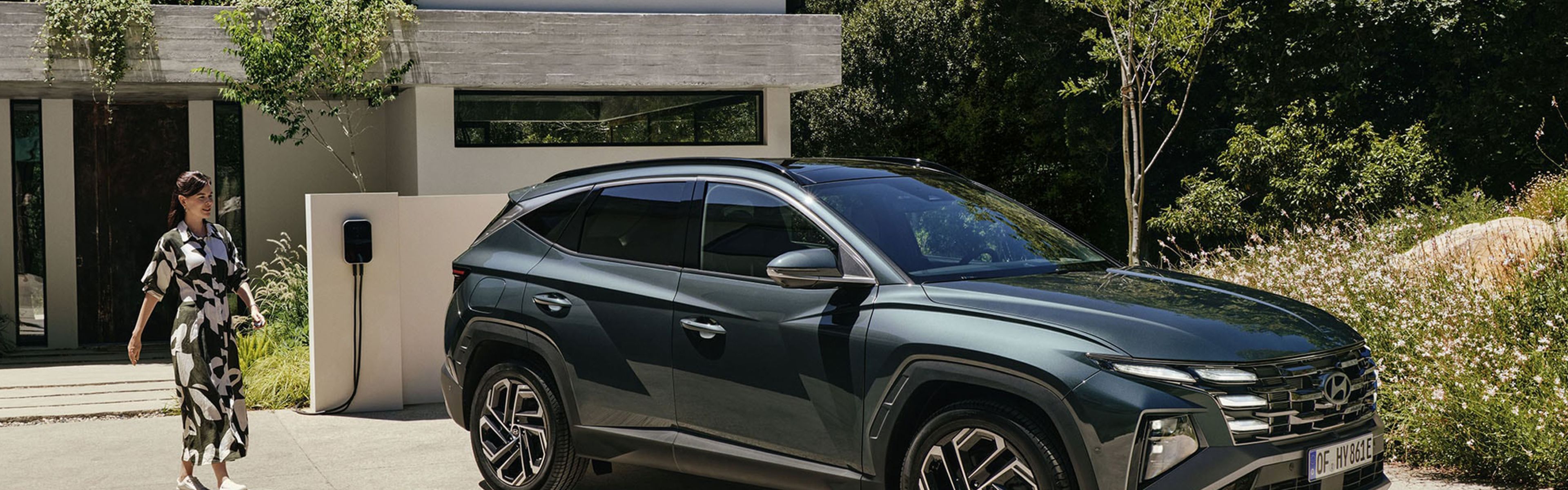 Der Hyundai TUCSON parkt vor einem modernen Haus mit Wallbox, eine Frau nähert sich von hinten dem Fahrzeug.