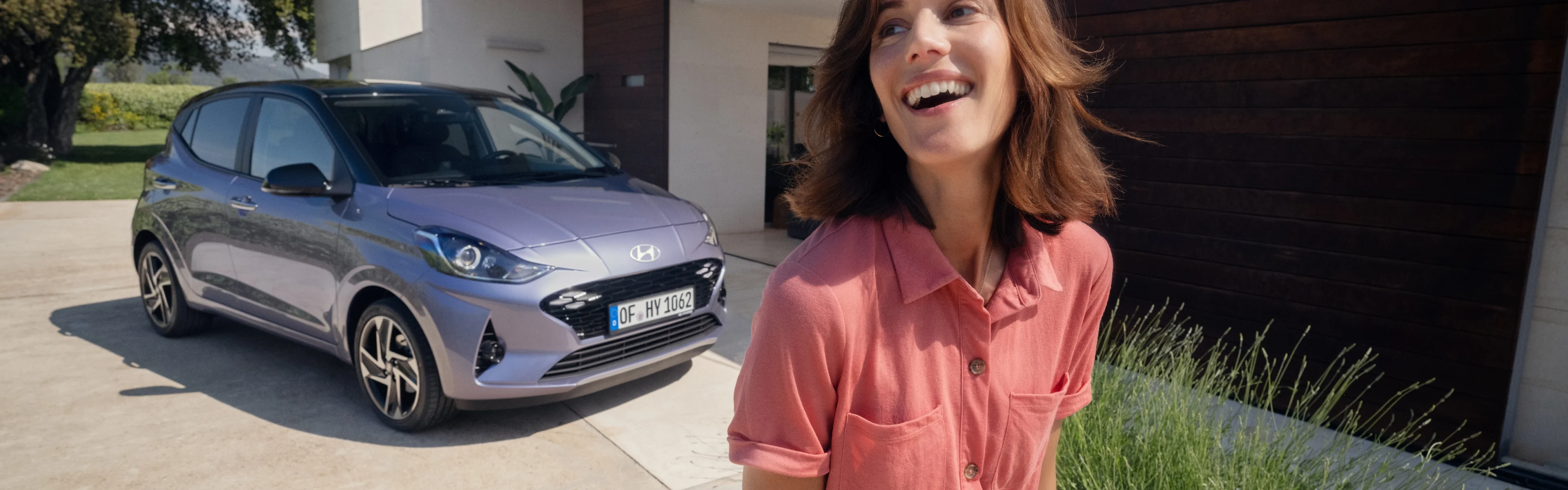 Ein Hyundai i10 parkt in der Einfahrt eines modernen Wohnhauses. Im Vordergrund lacht eine Frau in roter Bluse und Jeans.