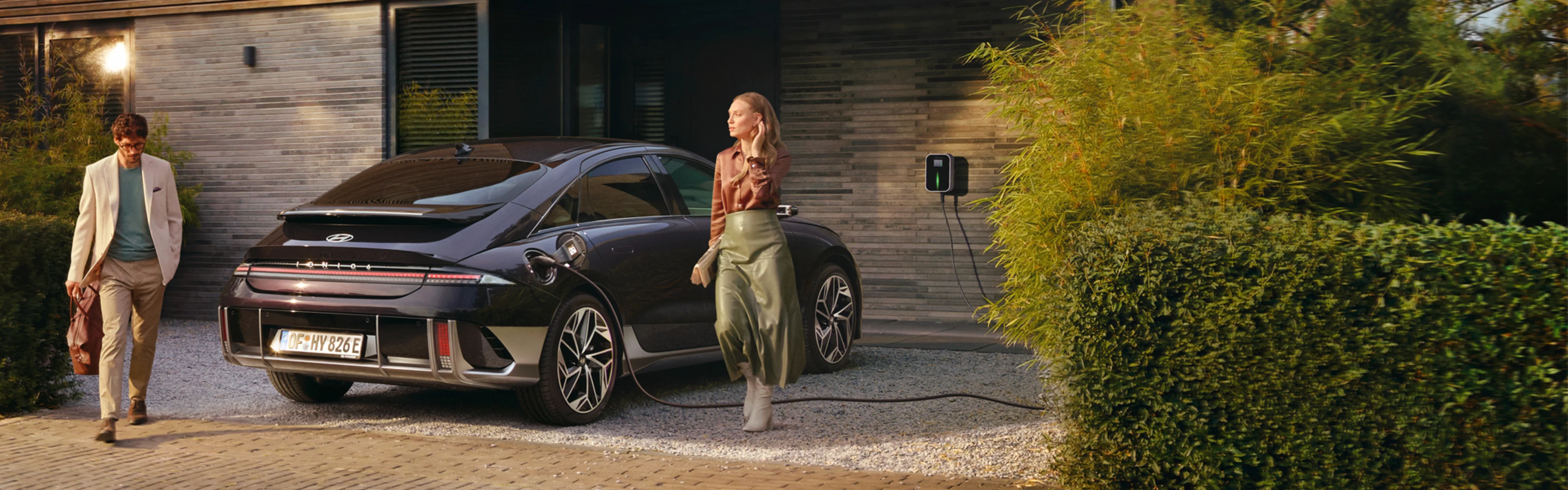 Ein schwarzer Hyundai IONIQ 6 ist an einer Wallbox vor einem modernen Haus angeschlossen, während eine Frau und ein Mann in eleganter Kleidung daneben stehen.