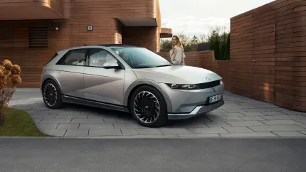 Eine Frau hinter ihrem Hyundai IONIQ 5 in der Einfahrt eines modernen, holzverkleideten Hauses.