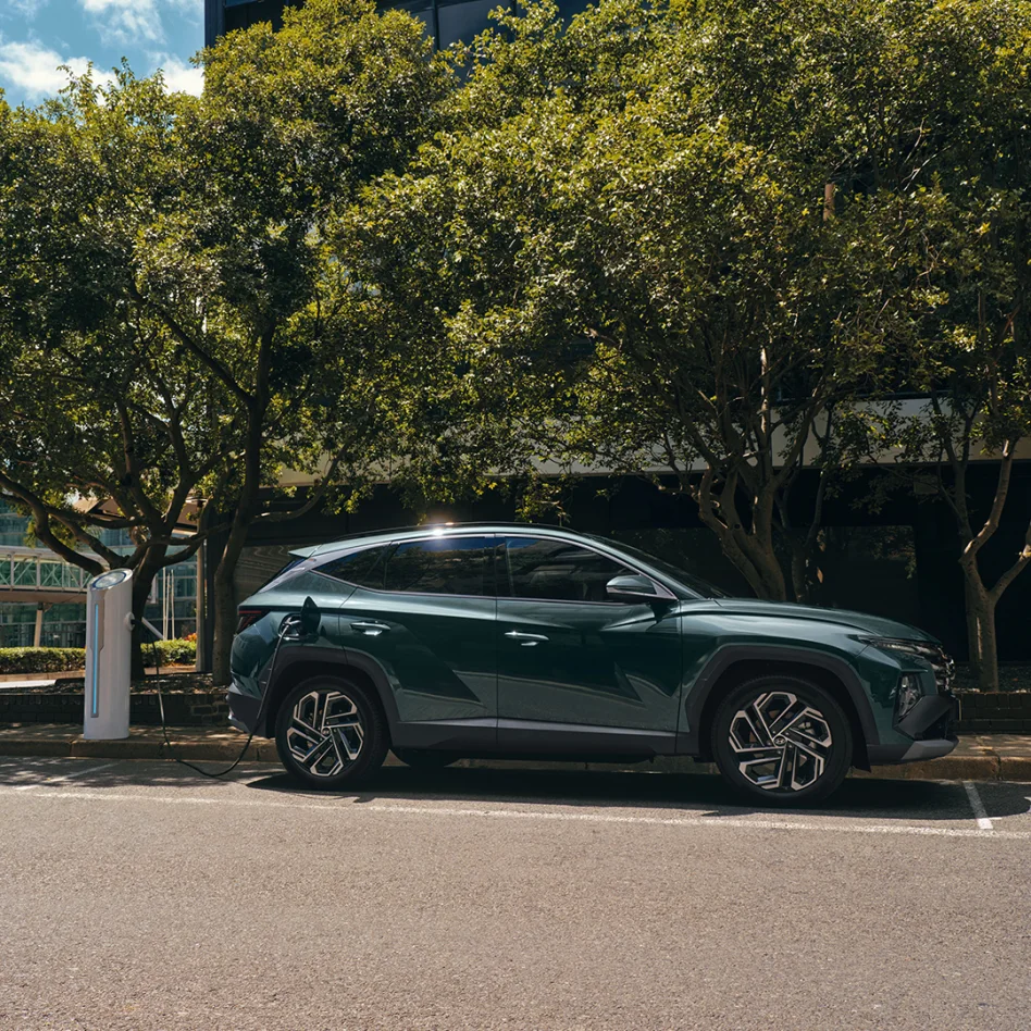 Dunkelgrüner Hyundai SUV lädt an einer öffentlichen Ladesäule unter Bäumen am Straßenrand.